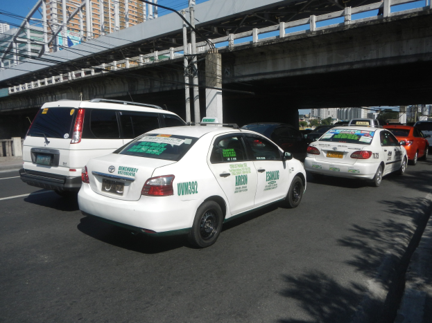 Eine Gruppe von Taxis steht neben einer Straße mit einer Brücke, Strommasten, Gebäuden, Bäumen und einem klaren blauen Himmel im Hintergrund.