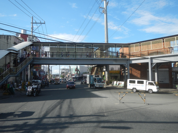 Stadtstraße mit Fahrzeugen, eine Fußgängerbrücke mit Menschen, Strommasten, Gebäude und eine bewölkte Himmel.