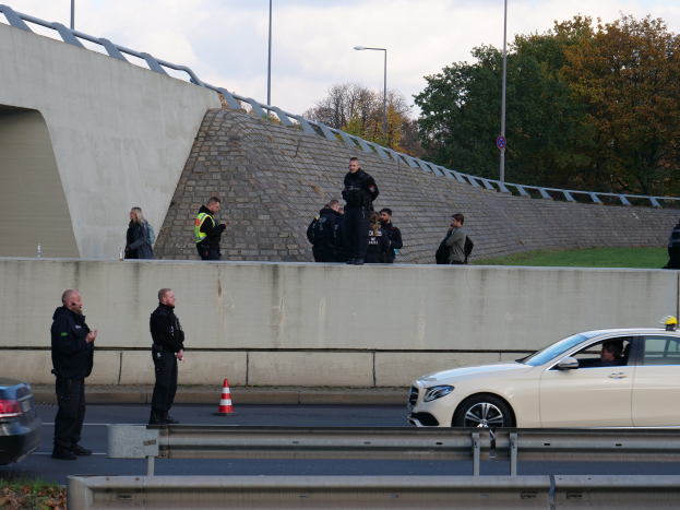 Eine Gruppe von Polizeibeamten steht an der Straße neben einem Auto, mit Verkehrskegeln, einer Trennwand, Gras, einer Wand, Laternenmästen, Bäumen und einem bewölktem Himmel im Hintergrund.