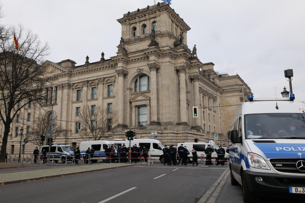 Eine Gruppe von Polizeibeamten steht vor dem Reichstagsgebäude in Berlin, Deutschland, mit Fahrzeugen, einem Zaun, Verkehrsampeln, Laternenmasten, Bäumen und Fahnen im Bild, bei klarem Himmel.