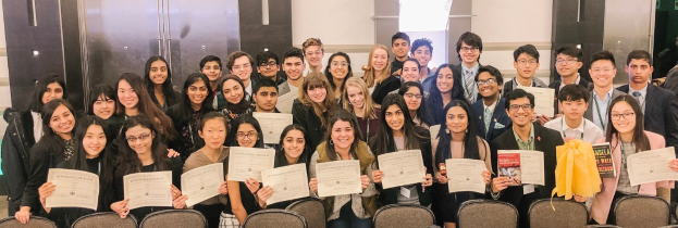 Gruppe von Menschen, die Zertifikate in einem Raum mit Stühlen und einer Wand im Hintergrund halten, wahrscheinlich bei einer Abschlussfeier für das akademische Jahr 2019-2020.