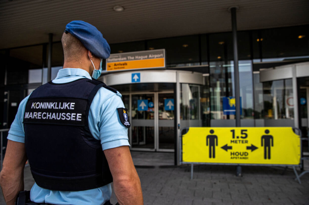 Ein Polizeibeamter in blauer Uniform und Mütze, der eine Maske trägt, steht vor einem Gebäude mit Glaswänden und einer gelben Tafel auf der rechten Seite sowie weiteren Tafeln im Hintergrund.