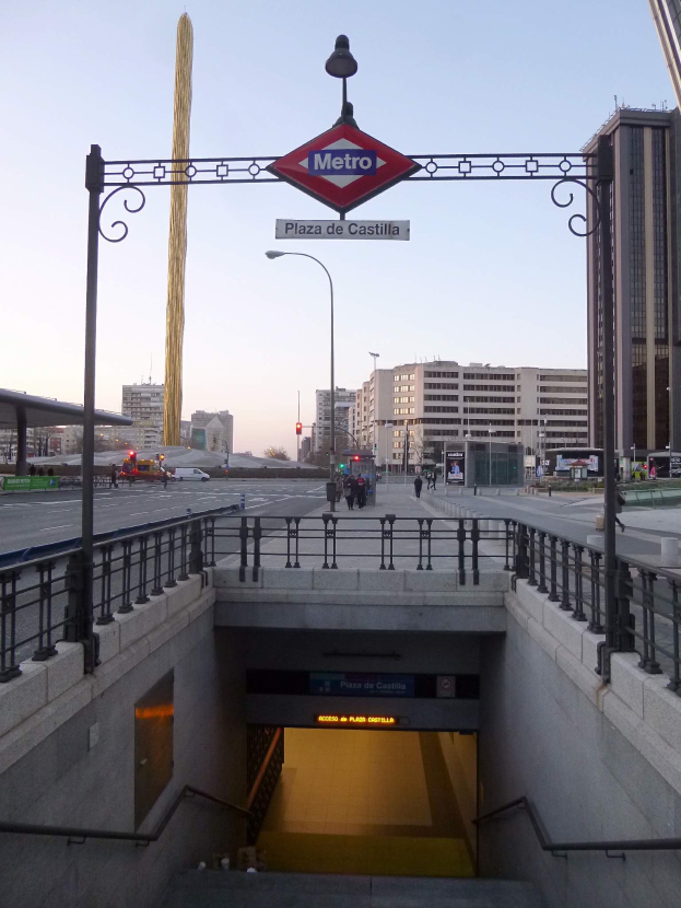 Eine belebte Stadtkulisse mit Gebäuden, Fahrzeugen, Fußgängern, einem U-Bahn-Eingang und einem Bogen mit einer Namensplakette im Hintergrund.