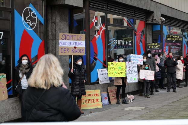 Eine Gruppe von Menschen mit Masken und Schildern protestiert vor einem Gebäude mit Glaswänden und -türen, wobei Tafeln mit Text und Logos sowie Kameras und ein Hund unten zu sehen sind.