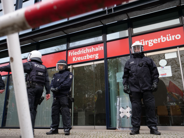 Polizeibeamte in Helmen und Handschuhen stehen vor einem Glasgebäude mit aufgeklebten Papieren und einem Pfahl links, während Schilder im Hintergrund "Friedrichshain Kreuzberg" anzeigen.