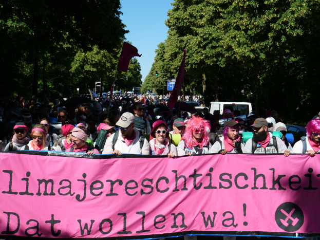 Eine Gruppe von Menschen marschiert mit einem pinken Banner, auf dem 'Women's March in Berlin' steht, durch die Straßen. Einige tragen Mützen und Schutzbrillen, andere halten Fahnen. Im Hintergrund sind Bäume und Fahrzeuge sowie ein klarer Himmel zu sehen.