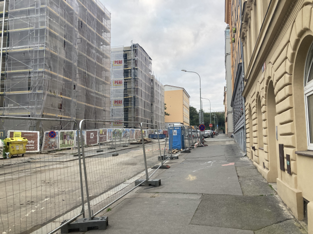 Eine Stadtstraße mit Gebäuden, Fahrzeugen und einer Baustelle mit Gerüsten im Vordergrund, unter einem bewölkten Himmel.