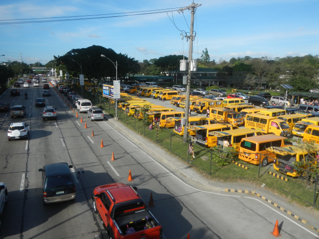 Eine belebte Straße mit zahlreichen gelben Schulbussen, Verkehrskegeln, Maschendrahtzaun, Laternenmasten, Strommasten mit Kabeln, Bäumen, Gebäuden und einem bewölkten Himmel im Hintergrund.