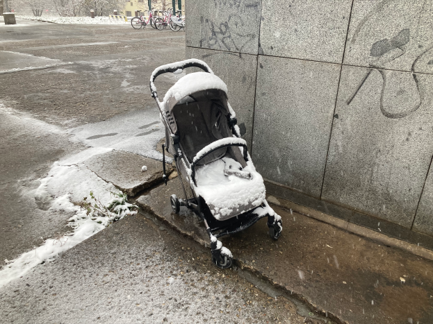 Ein Kinderwagen, der mit Schnee bedeckt ist, steht an der Seite einer Straße, mit einer Wand auf der rechten Seite des Bildes. Im Hintergrund gibt es Fahrräder, Bäume, Gebäude, Pfosten und einen Mülleimer, die alle von einer Schneeschicht bedeckt sind.