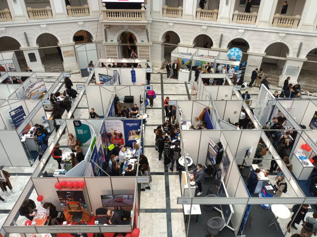 Eine große Gruppe von Menschen auf einer internationalen Messe mit Ständen, Bannern, Tischen, Stühlen und Ballons in einem Raum mit architektonischen Bögen, Säulen und einem Bildschirm im Hintergrund.