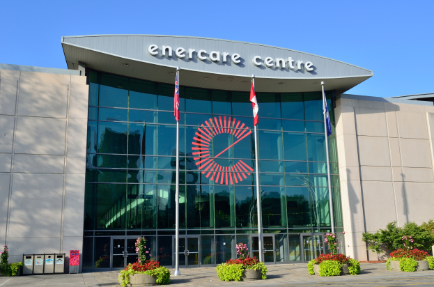 Ein großes Gebäude mit einem Schild, auf dem "enercare centre" steht, und Flaggen davor, umgeben von Pflanzen, Bäumen, Mülltonnen und einem Weg, unter einem bewölkten Himmel.