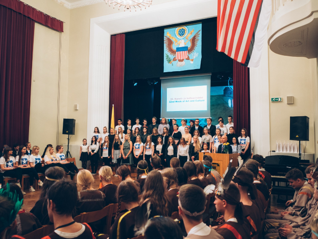 Eine Gruppe von Menschen, die vor einem sitzenden Publikum an einem Event stehen, mit einem Podium, Mikrofon, Flaggen, Lautsprechern, einem Bildschirm mit Text und einem Bild sowie Vorhängen und einem Kronleuchter im Hintergrund.