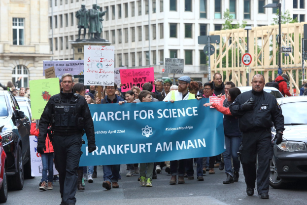 Gruppe von Menschen marschiert mit einem "March for Science Frankfurt am Main"-Schild die Straße entlang, Autos, Gebäude, Statuen, Laternenpfähle, Schilder und Bäume im Hintergrund.