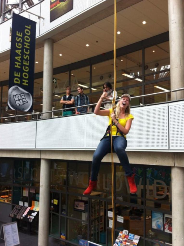 Eine Frau in einem gelben T-Shirt, einer Mütze und roten Stiefeln hängt an einem Seil in einem Buchladen, mit Regalen voller Bücher, einem Banner, ein paar Menschen, Glastüren und Deckenleuchten im Hintergrund.