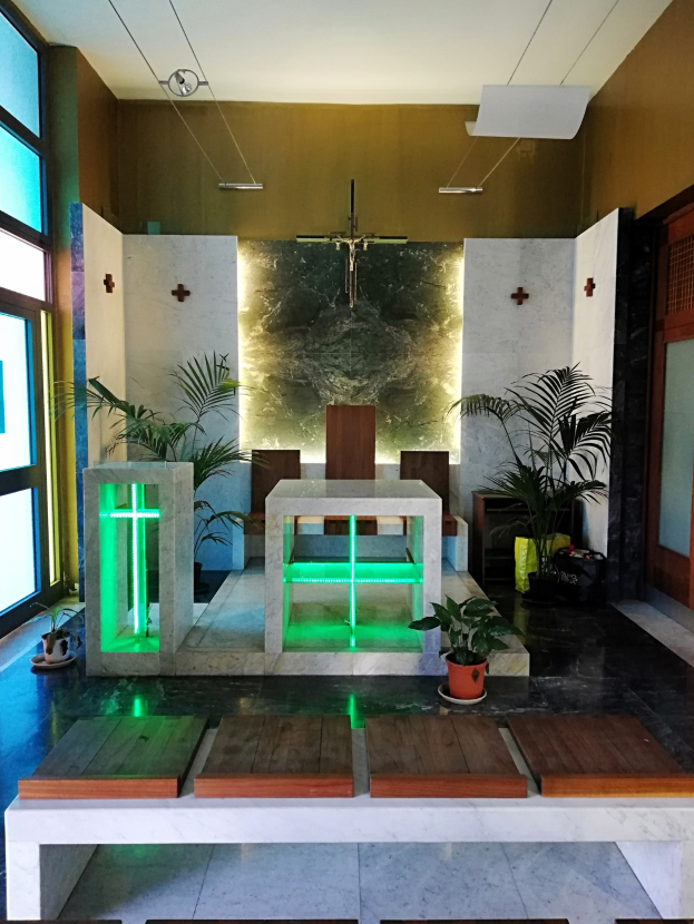 Innere einer Kirche mit Holzliegen, Zimmerpflanzen, einem Kreuz an der Wand, einer Tür auf der rechten Seite und Glasfenstern auf der linken Seite, beleuchtet von grünem Licht.