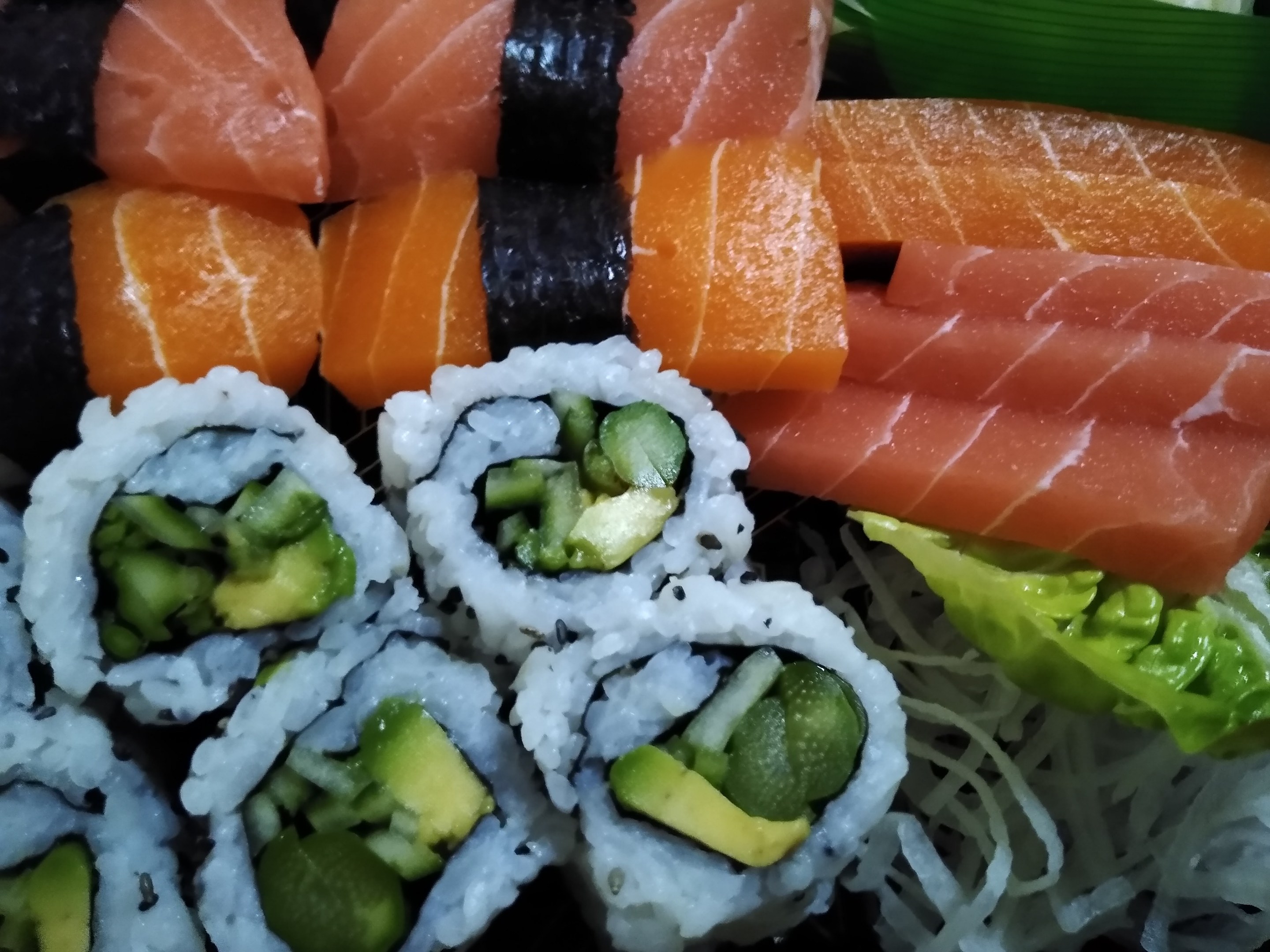 Ein schwarzer Teller mit verschiedenen Sushi-Stücken, darunter Lachs, Avocado und Gurke, umgeben von ein paar grünen Blättern.