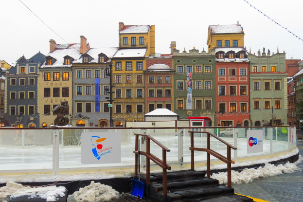 Eislaufbahn in einem Stadtplatz umgeben von Gebäuden, Treppen, Geländern, Informationsschildern, schneebedeckter Erde, Laternenmasten, Bäumen und Himmel.