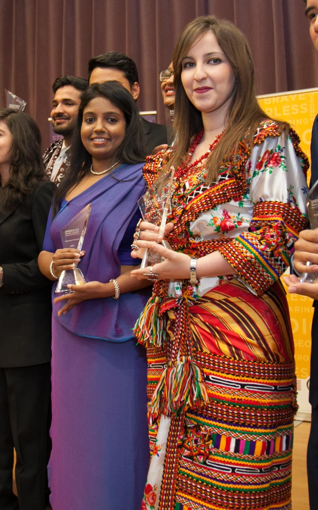 Eine Gruppe von Menschen steht auf einer Bühne mit einer Frau in der Mitte, die eine Trophäe hält, dahinter ist ein Vorhang und eine Tafel mit der Aufschrift "Bravo Segen Award Gewinner."
