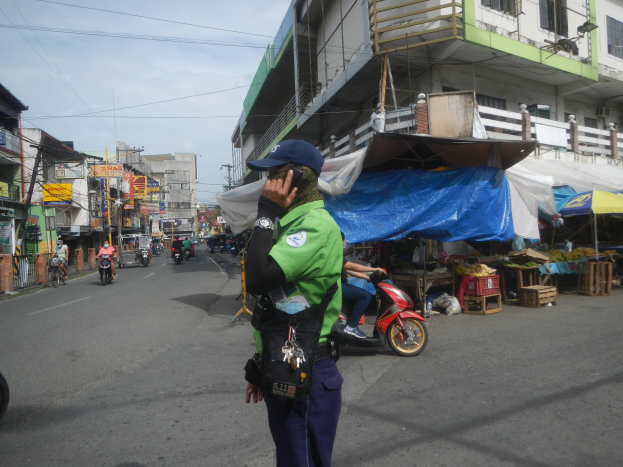 Ein Polizeibeamter in grüner und blauer Uniform und blauer Mütze steht an der Seite einer Straße, spricht auf einem Handy, mit Fahrzeugen im Hintergrund, Ständen mit Gegenständen zu seiner Rechten und einer bewölkten Himmel.