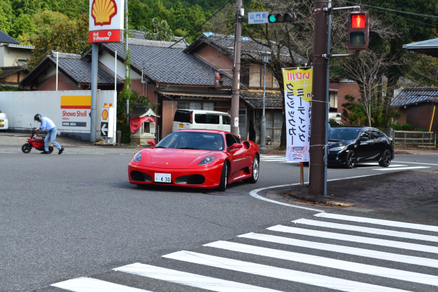 Roter Sportwagen fährt auf einer Straße neben einer Ampel, mit einer Person auf einem Roller im Vordergrund und Gebäuden, Bäumen und anderen Fahrzeugen im Hintergrund.