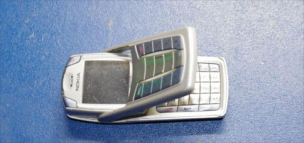 Eine Person, die strahlt und ein Mobiltelefon hält, während eine andere Person in der Nähe kauert, ein Rucksack, ein Megafon unten und eine Wand im Hintergrund, mit einer weiteren Person auf der linken Seite.