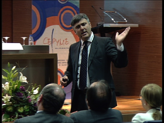 Ein Mann in einem schwarzen Blazer steht an einem Podium mit Mikrofonen, spricht in eines hinein, mit einem Blumentopf oder einem Bouquet in der Nähe und einem Tisch mit einem Buch, einer Wasserflasche und anderen Gegenständen daneben. Hinter ihm ist eine braune Wand und eine farbenfrohe Tafel mit Text zu sehen, was auf eine Konferenzhalle hindeutet.