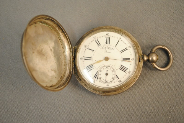 Eine silberne Taschenuhr mit einem weißen Zifferblatt mit römischen Zahlen, geschlossenem Deckel auf einer grauen Oberfläche.