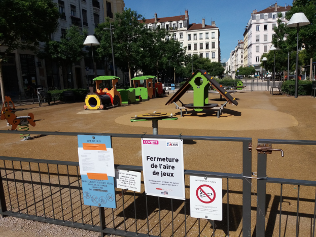 Umzäunter Spielplatz mit mehreren Schildern, umgeben von Bäumen, Laternenmasten und Gebäuden auf einem Stadtplatz unter einem sichtbaren Himmel.