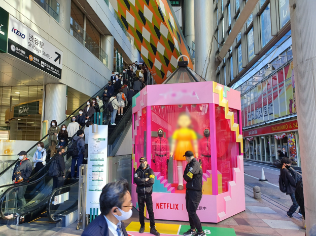 Eine Gruppe von Menschen steht vor einem gläsernen Gebäude mit Texttafeln, einige tragen Masken, während eine Rolltreppe mit Menschen zu sehen ist; ein pinker Kasten in der Mitte zeigt "Netflix's new Netflix booth in Tokyo" mit Schaufensterpuppen drinnen.