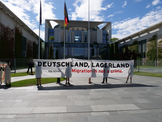 Eine Gruppe von Menschen mit einer Schilder mit der Aufschrift "Deutschland, Lagerland Migration ist kein Verbrechen" vor einem Gebäude mit Fenstern, umgeben von Gras, einem Metallzaun, Pflanzen, Bäumen und Fahnenmasten vor einem bewölkten Himmel.