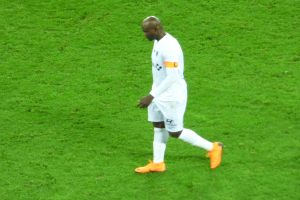 Ein Fussballspieler in einem wei├čen T-Shirt, wei├čen Shorts und orangefarbenen Schuhen ├╝berquert ein saftig gr├╝nes Feld.