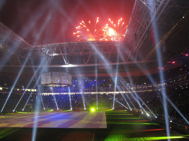 Das Olympische Stadion in London nachts, erleuchtet von hellen Lichtern und Feuerwerken, mit einem großen Bildschirm im Vordergrund und einer Menge drinnen.