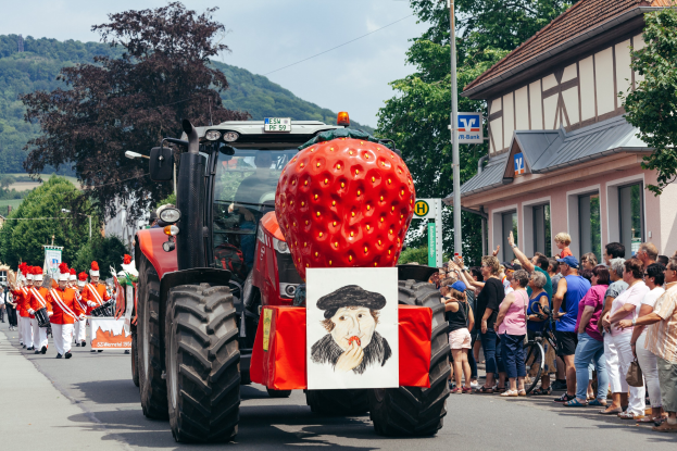 Ein großer Erdbeerförmiger Traktor fährt eine Straße entlang mit Menschen und Fahrrädern auf dem Gehweg, Gebäude und Bäume im Hintergrund unter einem klaren blauen Himmel.