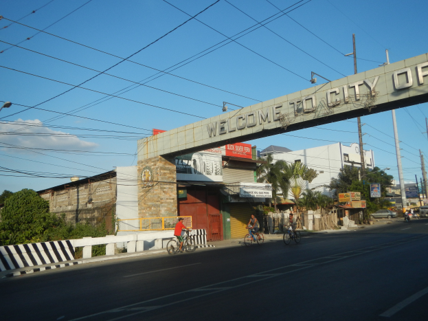 Eine Gruppe von Menschen, die auf Fahrr"{a}dern eine Stra{\ss}e in Manila entlangfahren, mit Geb"{a}uden, B"{a}umen und Strommasten an den Seiten, unter einem bew"{o}lktem Himmel und einem "{U}Willkommen in der Stadt Manila"-Bogen.