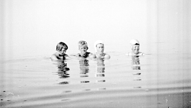 Vier Kinder in Schwarz-Weiß schwimmen in einem Pool, jedes trägt eine Schwimmkappe.