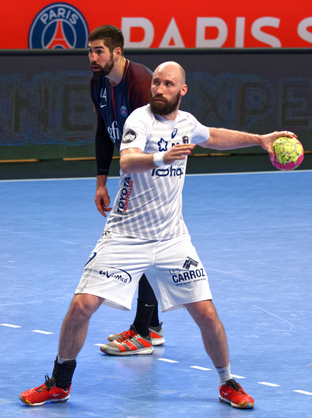 Zwei Männer spielen Handball auf einem Platz, einer hält einen Ball, im Hintergrund eine Tafel mit "Paris Saint-Germain vs Paris Saint Germain."