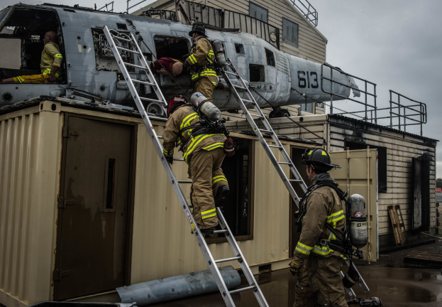 Feuerwehrleute in Uniform und Helmen arbeiten an einem Hubschrauber auf einem Gebäudedach, einer hält einen Sauerstoffzylinder, mit Leitern, einem Container, Bäumen und einem klaren blauen Himmel im Hintergrund.