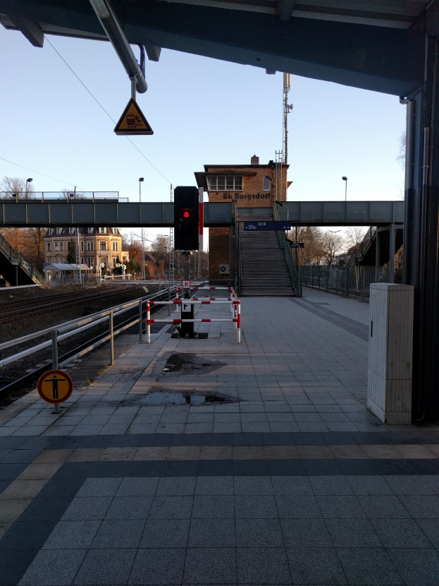 Ein Bahnsteig mit einem Zug auf den Schienen, eine Plattform mit Geländern, ein Schild, eine Verkehrsampel an einem Pfahl, eine Brücke mit Treppen, Laternenpfähle, Bäume, Gebäude mit Fenstern, ein Turm und ein Himmel im Hintergrund.