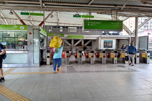 Menschen gehen durch einen Bahnhof mit Metalldetektoren, Hinweistafeln, Metallstangen, Deckenleuchten und Gebäuden mit Fenstern im Hintergrund.