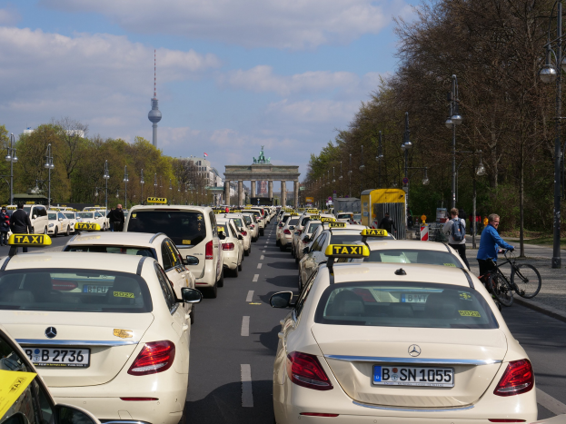 Eine lange Reihe von Taxis, die entlang einer belebten Straße in Berlin, Deutschland, geparkt sind, mit Fahrzeugen, Fahrradfahrern und Fußgängern in der Nähe, flankiert von Laternenmasten, Bäumen und Gebäuden, einschließlich eines Bogens und eines Turms.