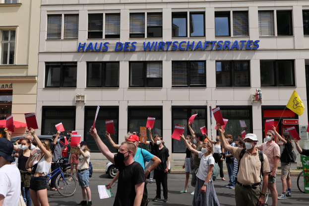 Eine Gruppe von Menschen protestiert vor einem Gebäude, hält Fahnen und Schilder hoch, mit Fahrzeugen auf der Straße und einem Schild auf der rechten Seite.