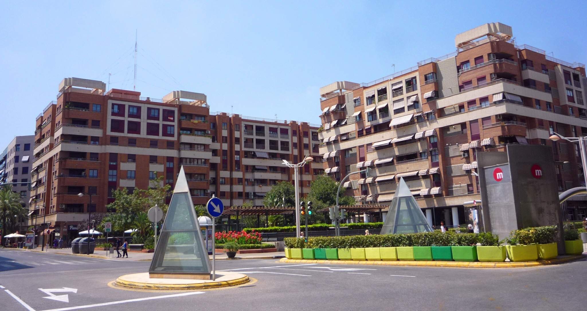 Eine Stadtstraße mit einem zentralen Denkmal, umgeben von hohen Gebäuden, Straßenmöbeln, Grünflächen, Fußgängern und einem klaren blauen Himmel.