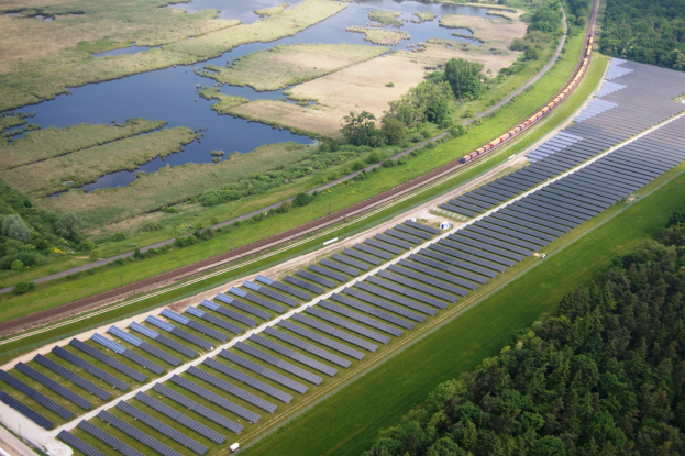 Luftaufnahme einer Solarpark mit Panelen, umgeben von Bäumen, Gras, Wasser und einem Zug auf einer nahen Bahnstrecke.