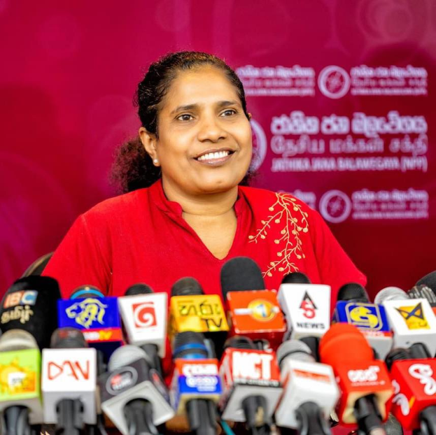 Eine Frau in einem roten Hemd sitzt an einem Tisch mit Mikrofonen, lächelt selbstbewusst und hat ein Banner im Hintergrund mit der Aufschrift "Sri Lankas Ministerin für Gesundheit und Soziales".