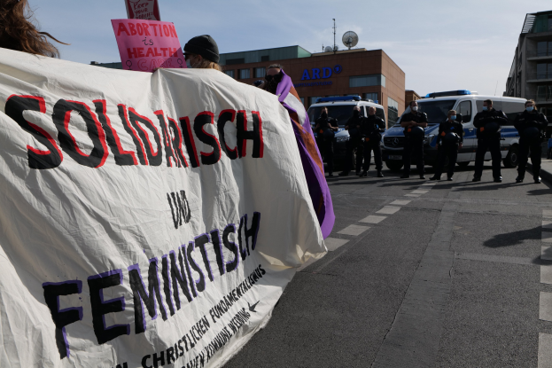 Gruppe von Menschen, die eine Straße entlanggehen und eine 'Solidarität und Feminismus'-Schleife halten, mit parkenden Fahrzeugen, Gebäuden, einer Schüsselantenne und einem klaren blauen Himmel im Hintergrund.