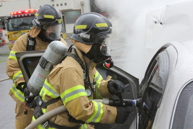 Feuerwehrleute in Schutzausrüstung löschen ein brennendes Auto mit einem Schlauch, während Rauch daraus aufsteigt, mit Fahrzeugen und einem Gebäude im Hintergrund.