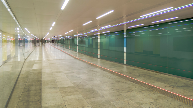 Ein langer Gang mit Glaswänden auf beiden Seiten und einer beleuchteten Decke, mit ein paar Menschen, die einen belebten Eindruck einer Stadtlandschaft erwecken.