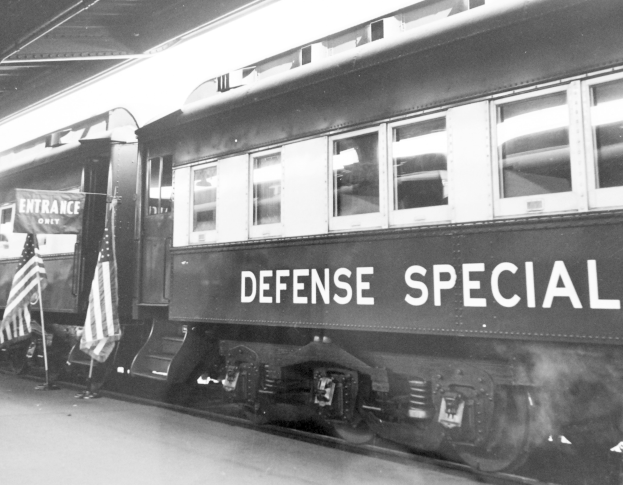 Schwarz-weißes Foto eines Zugs mit der Aufschrift "Defense Special" auf den Eisenbahnschienen mit zwei Fahnen links und einer Decke oben.