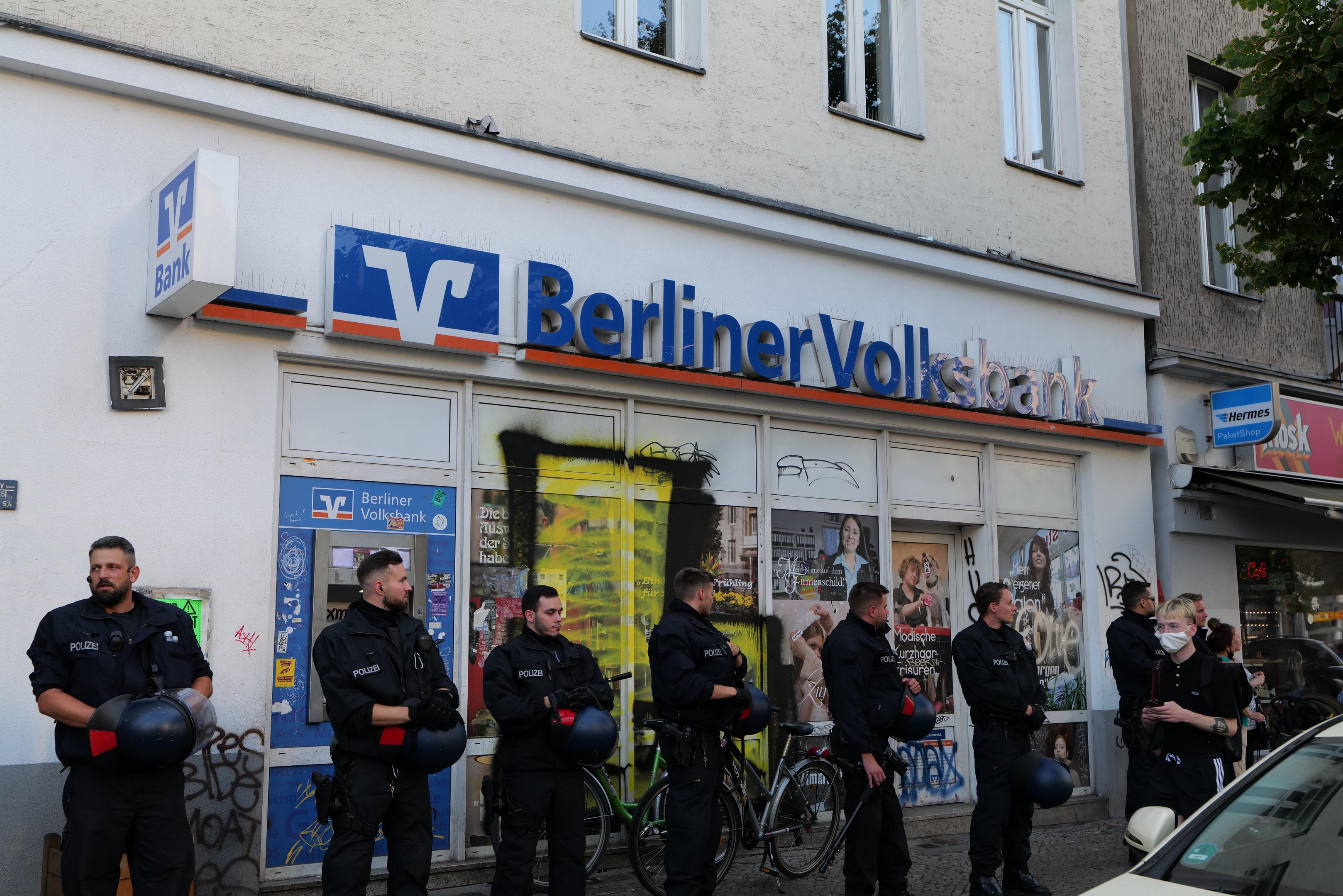 Gruppe von Polizisten vor einem Berliner Volksbank-Gebäude stehend, einige halten Helme, mit einem Auto rechts und einem Baum links, Gebäude mit Fenstern und Texttafeln im Hintergrund.