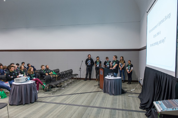 Eine Person, die in ein Mikrofon spricht, bei einer Versammlung mit sitzenden Zuschauern, Mannschaftsmitgliedern mit Ausweisen, einem Präsentationsbildschirm und einem Videoabspieler in einem leeren Büroraum.